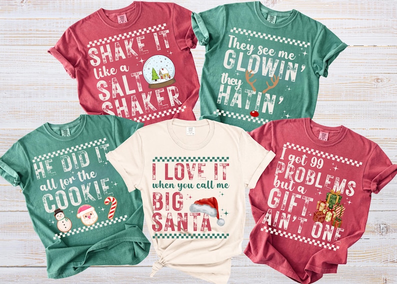 Op de afbeelding: Een verzameling van vijf t-shirts met korte mouwen in verschillende kleuren, elk met een kerstthema en tekst. De shirts zijn rood, groen en cr&egrave;mekleurig, met zinnen als "Shake it like a salt shaker" en "I love it when you call me Big Santa".