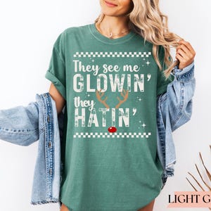 Op de afbeelding: Lichtgroen T-shirt met de witte tekst "They see me glowin', they hatin'". Het ontwerp bevat rendiergeweien en een rode neus. Het shirt heeft een relaxte pasvorm.