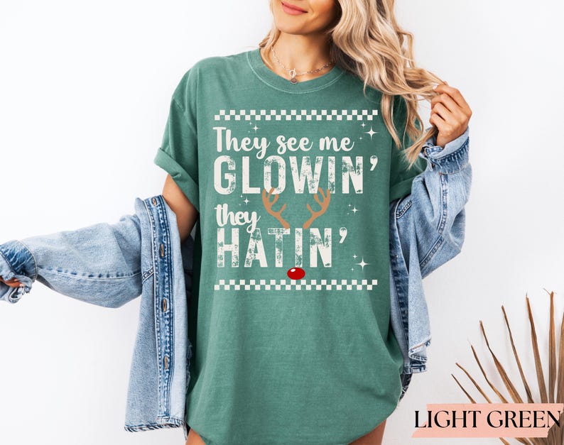 Op de afbeelding: Lichtgroen T-shirt met de witte tekst "They see me glowin', they hatin'". Het ontwerp bevat rendiergeweien en een rode neus. Het shirt heeft een relaxte pasvorm.