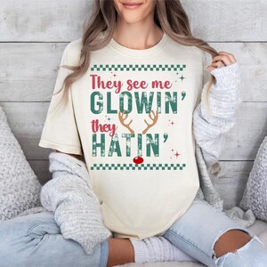 Op de afbeelding: Cr&egrave;mekleurig T-shirt met de tekst "They see me GLOWIN' they HATIN'" in rood en groen. Het ontwerp bevat rendiergeweien en een rode neus, met een groene geblokte rand. Het shirt is een casual, feestelijk ontwerp.