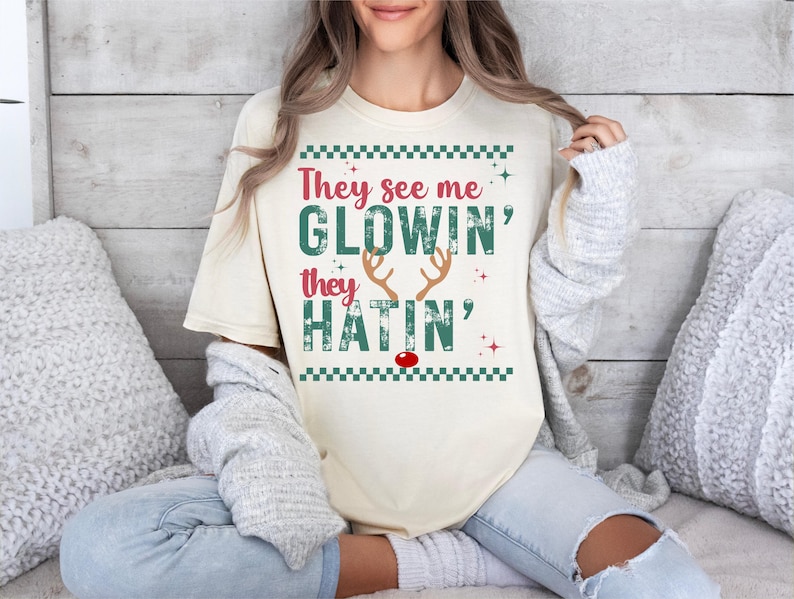 Op de afbeelding: Cr&egrave;mekleurig T-shirt met de tekst "They see me GLOWIN' they HATIN'" in rood en groen. Het ontwerp bevat rendiergeweien en een rode neus, met een groene geblokte rand. Het shirt is een casual, feestelijk ontwerp.