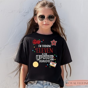 May include: Black t-shirt with the text "I'm turning eleven in the upside down" and various graphics. The graphics include a waffle, a cassette tape, a star, and dice. The shirt is worn by a person wearing heart-shaped sunglasses.