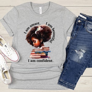 May include: Gray t-shirt with a graphic of a Black girl with an afro, reading a stack of books. The text on the shirt reads "I am smart. I am beautiful. I am confident."