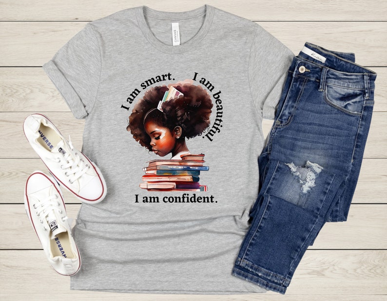 May include: Gray t-shirt with a graphic of a Black girl with an afro, reading a stack of books. The text on the shirt reads "I am smart. I am beautiful. I am confident."