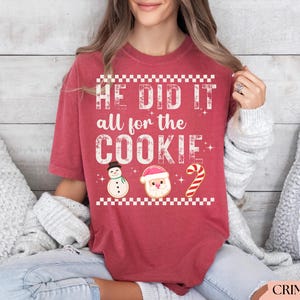 Op de afbeelding: Karmizijnrood T-shirt met de tekst "HE DID IT all for the COOKIE". Het shirt heeft koekjesontwerpen van een sneeuwpop, de kerstman en een zuurstok. Het shirt heeft een relaxte pasvorm.