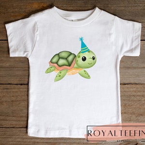 Pu&ograve; includere: Maglietta bianca con una tartaruga verde e arancione che indossa un cappello da festa blu e giallo. La tartaruga sorride e ha occhi grandi. La maglietta ha il testo "ROYAL TEEFINDS APPAREL" in basso.
