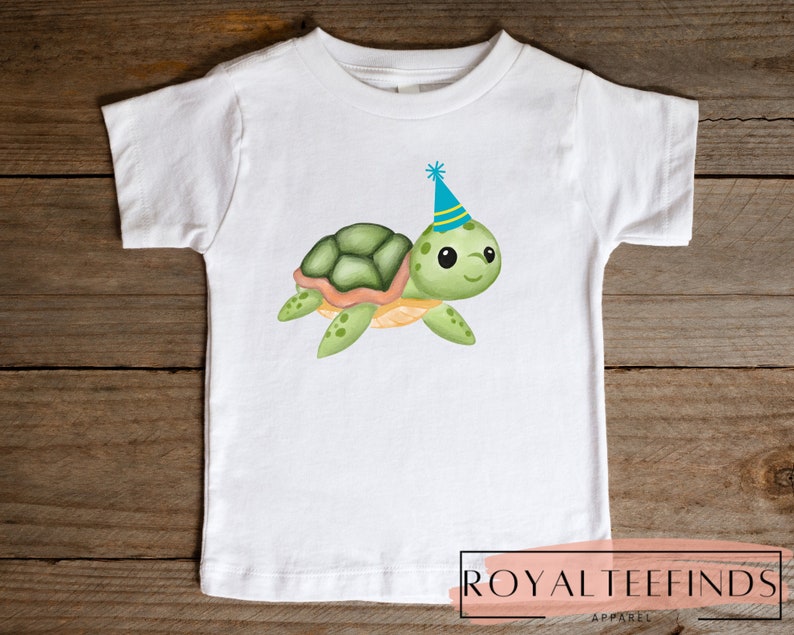 Pu&ograve; includere: Maglietta bianca con una tartaruga verde e arancione che indossa un cappello da festa blu e giallo. La tartaruga sorride e ha occhi grandi. La maglietta ha il testo "ROYAL TEEFINDS APPAREL" in basso.