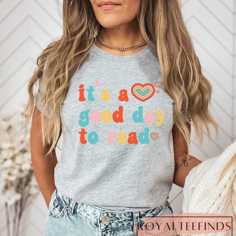 K&ouml;nnte beinhalten: Ein graues T-Shirt mit dem Text "it's a good day to read" in bunten Buchstaben mit einem Regenbogenherz-Motiv.