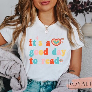 K&ouml;nnte beinhalten: Wei&szlig;es T-Shirt mit dem Text "it's a good day to read" in bunter Retro-Schrift mit Herzen.