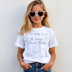May include: A white t-shirt with the text "The Summer I Turned Thirteen" in light blue, along with a seashell graphic. The shirt is worn by a person wearing pink heart-shaped sunglasses and blue jeans.