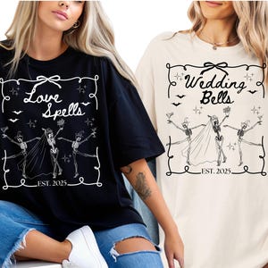 May include: Black and cream-colored t-shirts with a graphic design featuring skeleton figures in wedding attire. The shirts have the phrases "Love Spells" and "Wedding Bells" with the year "EST. 2025" and decorative elements.