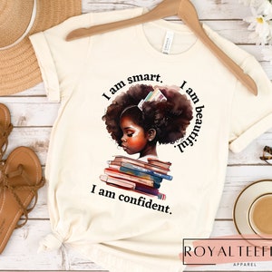 May include: A white t-shirt with a graphic of a young Black girl with a stack of books. The text on the shirt reads "I am smart. I am beautiful. I am confident."