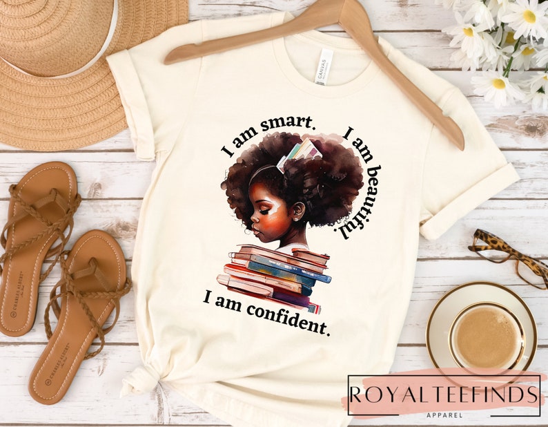 May include: A white t-shirt with a graphic of a young Black girl with a stack of books. The text on the shirt reads "I am smart. I am beautiful. I am confident."