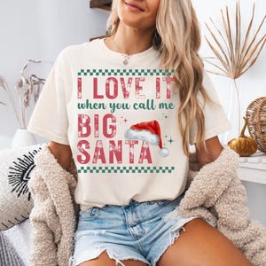 Op de afbeelding: Cr&egrave;mekleurig T-shirt met de tekst "I LOVE IT when you call me BIG SANTA" in rode letters. Het ontwerp bevat een kerstmuts en groene geblokte randen, perfect voor de feestdagen.