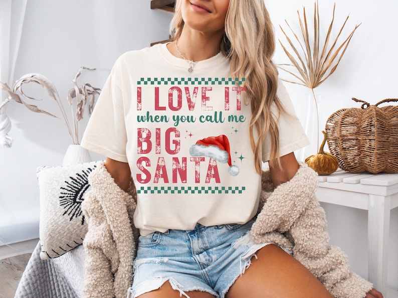 Op de afbeelding: Cr&egrave;mekleurig T-shirt met de tekst "I LOVE IT when you call me BIG SANTA" in rode letters. Het ontwerp bevat een kerstmuts en groene geblokte randen, perfect voor de feestdagen.