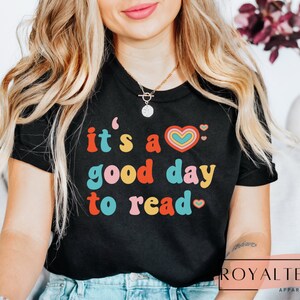 K&ouml;nnte beinhalten: Ein schwarzes T-Shirt mit dem Text "it's a good day to read" in bunter Retro-Schrift. Neben dem Text befindet sich eine Regenbogenherz-Grafik.