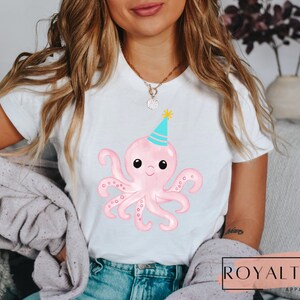 Pu&ograve; includere: Una maglietta bianca con un polpo rosa che indossa un cappello da festa blu e turchese. Il polpo ha occhi neri e una bocca sorridente.
