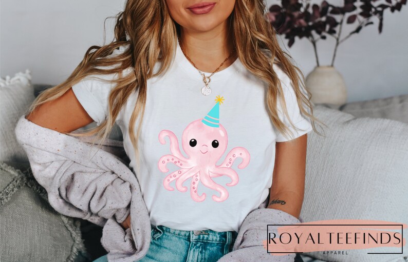 Pu&ograve; includere: Una maglietta bianca con un polpo rosa che indossa un cappello da festa blu e turchese. Il polpo ha occhi neri e una bocca sorridente.