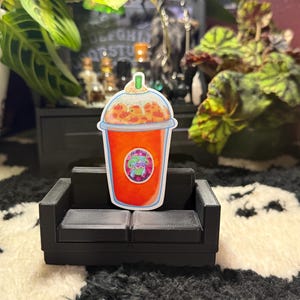 May include: A sticker of a colorful iced drink with whipped cream and a green straw, placed on a miniature black couch. The drink has an orange base and a green and purple logo. The background includes plants and a black cabinet.