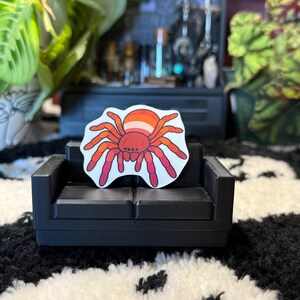 May include: A sticker of a spider in the colors of the lesbian pride flag rests on a miniature black couch. The spider has a red body and legs, with orange and pink accents. The couch is placed on a black and white patterned surface.