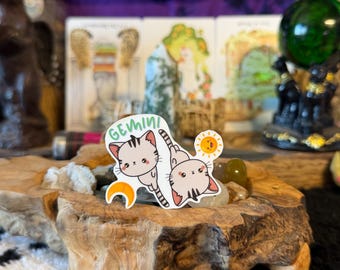 May include: A die-cut sticker featuring two light beige cartoon cats with dark stripes, positioned back-to-back. The word GEMINI is printed in green above the cats, accompanied by a small yellow sun icon and an orange crescent moon icon.