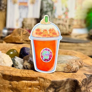 May include: A die-cut sticker of an orange beverage in a clear plastic cup with a dome lid and green straw. The drink is topped with whipped cream and small orange pumpkin shapes. A circular logo with a green-haired figure is on the cup front.