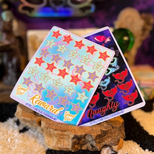 May include: Two rectangular sticker sheets featuring colorful designs. The left sheet has a light blue background with rows of pink and purple stars labeled Good Girl and the text Good Girl Reward. The right sheet has a dark background with red devil-themed Bad Girl stickers and the text Naughty.
