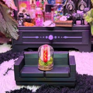 May include: A miniature hot dog in a glass dome sits on a small, dark green couch. The hot dog has a red sausage, yellow mustard, and green relish. The couch is placed on a black and white patterned rug, with a black cabinet in the background.