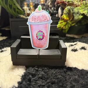 May include: A colorful sticker of a pink drink with whipped cream and a green straw, placed on a miniature black couch. The drink has a logo with a green creature. The background includes plants and a black and white rug.