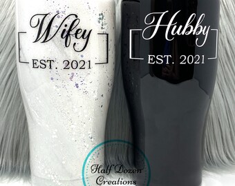 May include: Two insulated tumblers, one white with a marble effect and the word "Wifey" and "EST. 2021", the other black with "Hubby" and "EST. 2021". Both tumblers have black lids and are from Half Dozen Creations.