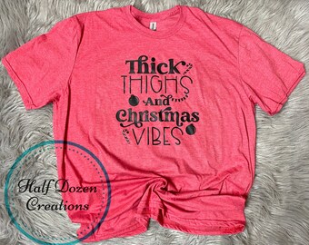 May include: A red t-shirt with the text "Thick Thighs And Christmas Vibes" in black. The design includes Christmas ornaments and candy canes. The shirt also features the text "Half Dozen Creations" in a blue circle.