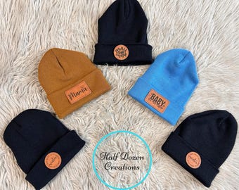 May include: Five beanies in various colors: black, blue, and tan. Each beanie has a leather patch with text, including "Mama", "Baby", and "Winter". The beanies are displayed on a fluffy, white surface.