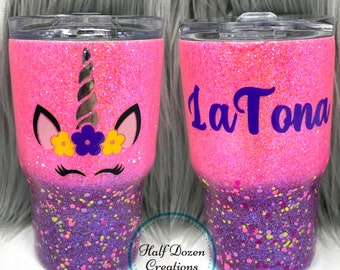 May include: Two glitter-covered tumblers. One features a unicorn face with a silver horn, black ears, and floral accents. The other has the name "LaTona" in purple script. The tumblers transition from pink to purple with colorful speckles.