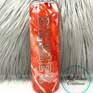 May include: A stainless steel tumbler with a red and white marbled design. The word "DREZZEK" is vertically etched in silver. The tumbler has a clear lid and is cylindrical in shape. The tumbler is a drinkware item.