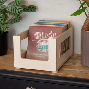 May include: A wooden record crate with a stack of vinyl records inside. The crate is light brown and has a simple design. The records are various colors and have different titles. The crate is sitting on a wooden surface with a black drawer underneath.