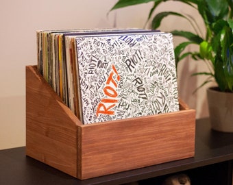 May include: A wooden record storage box with a stack of vinyl records inside. The records have a black and white design with the word "RIOT!" written in orange. The box is sitting on a black and wood countertop.