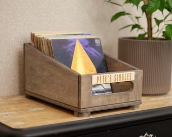May include: A wooden record crate with a label that reads "Pete's Singles" and is filled with vinyl records. The crate is sitting on a wooden surface next to a potted plant.