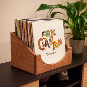 Puede incluir: Soporte de discos de madera lleno de discos de vinilo. La portada del álbum visible presenta el texto "Eric Clapton Behind the Sun" en letras coloridas. El soporte está sobre una superficie oscura con una planta en el fondo.