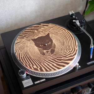 Könnte beinhalten: Schwarzer Plattenspieler mit einer individuellen Slipmat zum Thema Katze. Die Slipmat zeigt ein braunes Katzendesign in der Mitte, umgeben von konzentrischen Wirbelmustern. Der Plattenspieler steht auf einem schwarzen Stereoschränkchen.