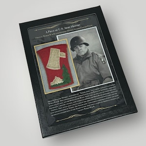 May include: A framed display of a World War II 70th Infantry Division soldier's uniform patch. The patch is red, white, and green with a stylized axe and pine tree. The text reads "A Piece of U.S. Army History."  The 70th Infantry Division, nicknamed the "Trailblazers," was a highly effective unit during World War II. They participated in key campaigns such as the Battle of Normandy, liberating towns and villages, and the Battle of the Bulge, where they helped stop the enemy offensive. The division's bravery and skill were evident in crossing the Rhine River and pushing deep into Germany. They also played a crucial role in the liberation of concentration camps and their post-war occupation of Europe. The Trailblazers made a significant contribution to the Allied victory in World War II.