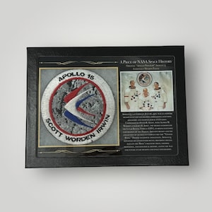 Könnte beinhalten: Ein gerahmtes Schwarzweiß-Apollo-15-Missionsabzeichen mit dem Text "Apollo 15" und den Namen "Scott, Worden, Irwin". Das Abzeichen zeigt einen blauen und roten Vogel, der über einer grauen und weißen Mondoberfläche fliegt. Der Rahmen enthält eine Beschreibung der Apollo-15-Mission.