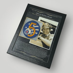 Op de afbeelding: Een ingelijst display van een vintage World War II 5th Air Force pilot uniform patch. De patch is blauw met een geel nummer 5 en een rode ster. De lijst bevat een zwart-witfoto van een glimlachende piloot die de patch draagt.