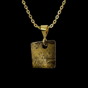May include: A gold-colored necklace with a square pendant. The pendant has a textured surface and a small hole near the top. The chain and pendant are a matching gold color, set against a black background.