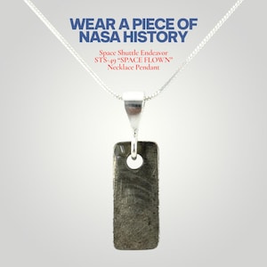 Puede incluir: Un collar de plata con un colgante rectangular. El colgante es de color gris oscuro con un agujero circular cerca de la parte superior. El texto "WEAR A PIECE OF NASA HISTORY" está en la parte superior, con "Space Shuttle Endeavor STS-49 'SPACE FLOWN' Necklace Pendant" debajo.