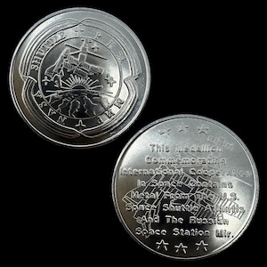 以下が含まれることがあります： 2つの銀色の記念コイン。1つのコインにはスペースシャトル、NASAのロゴ、そして「MIR」の文字が描かれています。もう1つのコインには「This Medallion Commemorating International Cooperation In Space Contains Metal From the U.S. Space Shuttle Atlantis And The Russian Space Station Mir.」と書かれています。