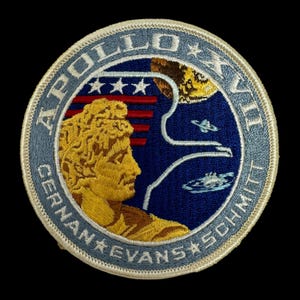 May include: A blue and white embroidered patch with the text "Apollo XVII, Cernan, Evans, Schmitt" and a depiction of a man's head with a moon and stars in the background.