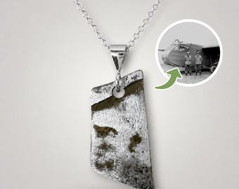May include: A silver-toned pendant necklace featuring an irregular, weathered metal fragment with dark, textured patches. A circular inset shows a black and white historical photograph of two individuals standing in front of a large aircraft.