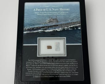 Pode incluir: Exibição emoldurada intitulada "A Piece of U.S. Navy History" com uma fotografia do porta-aviões USS Enterprise. Inclui um pequeno fragmento de madeira e um emblema prateado, comemorando o serviço do navio na Segunda Guerra Mundial.