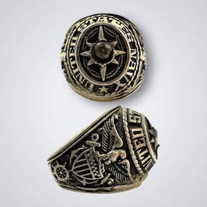 May include: Two views of a silver-toned ring. The top view shows the ring's face with the words "UNITED STATES NAVY" around a central starburst design. The side view reveals detailed nautical and eagle motifs.