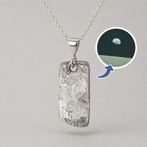 May include: A silver-colored pendant necklace with a rectangular, textured pendant. The pendant hangs from a delicate chain. A small image of the Earth from space is in the upper right corner. The pendant has a hole at the top for the chain.